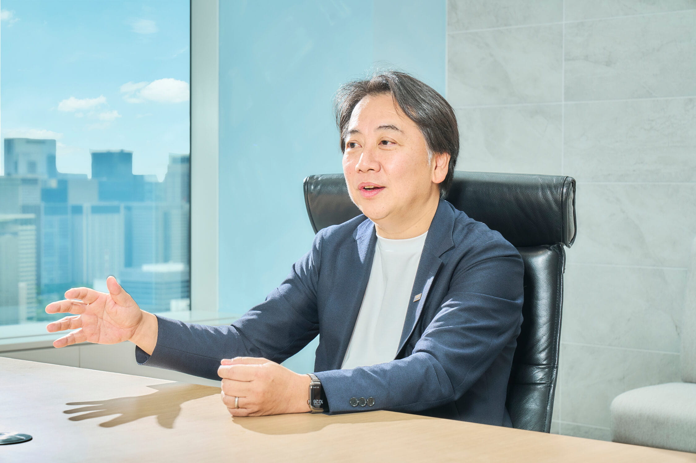 都市を背景にデスクで語る代表取締役社長 落合孝之